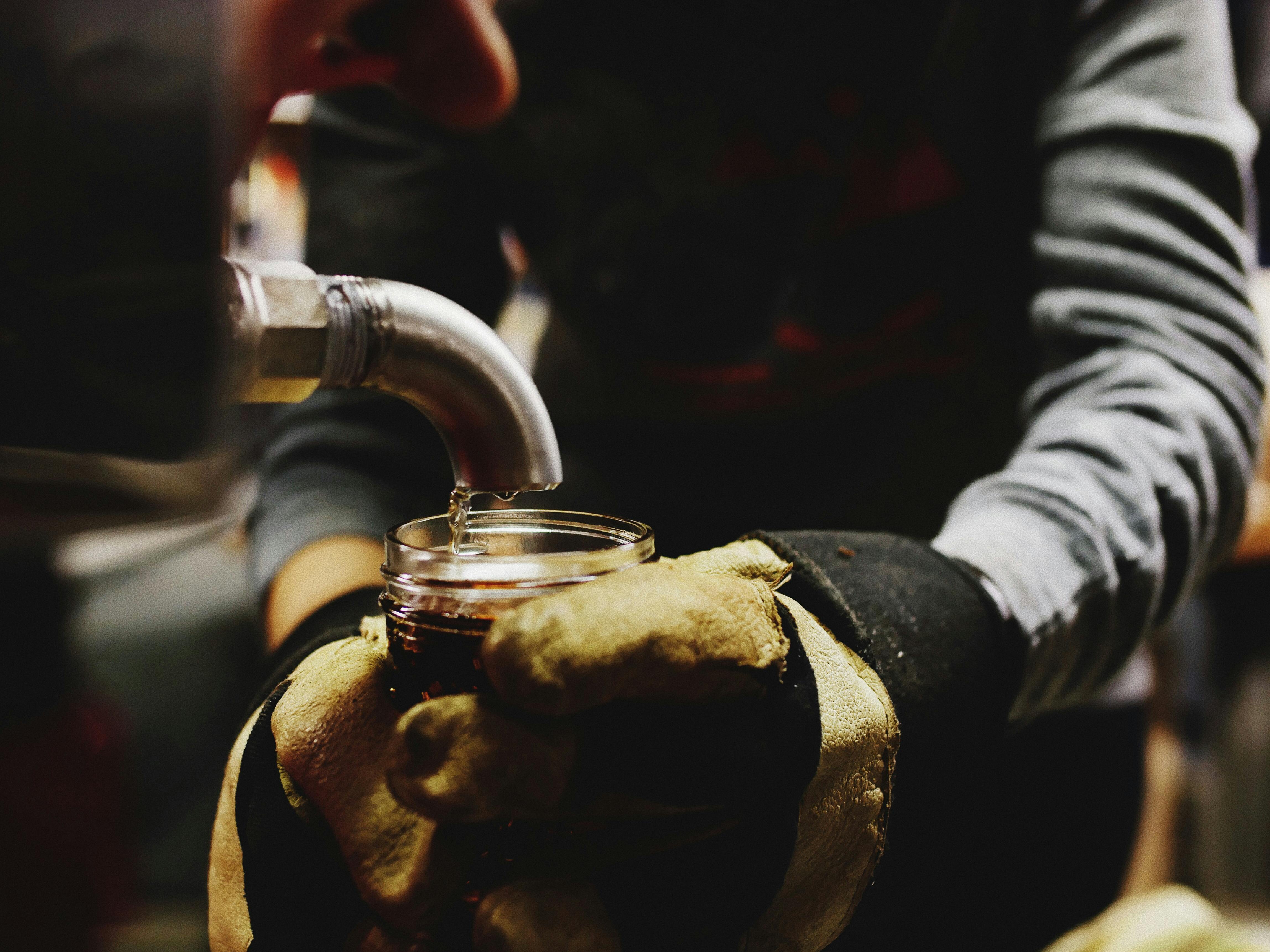 Person wearing gloves holding a jar and filling up with maple syrup from a tap.